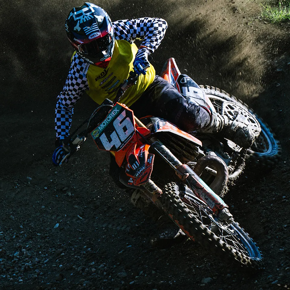 En motocrossfører i en gul jakke med svart-hvite sjakketærmer og en hjelm kjører en oransje motorsykkel på en støvete bane.